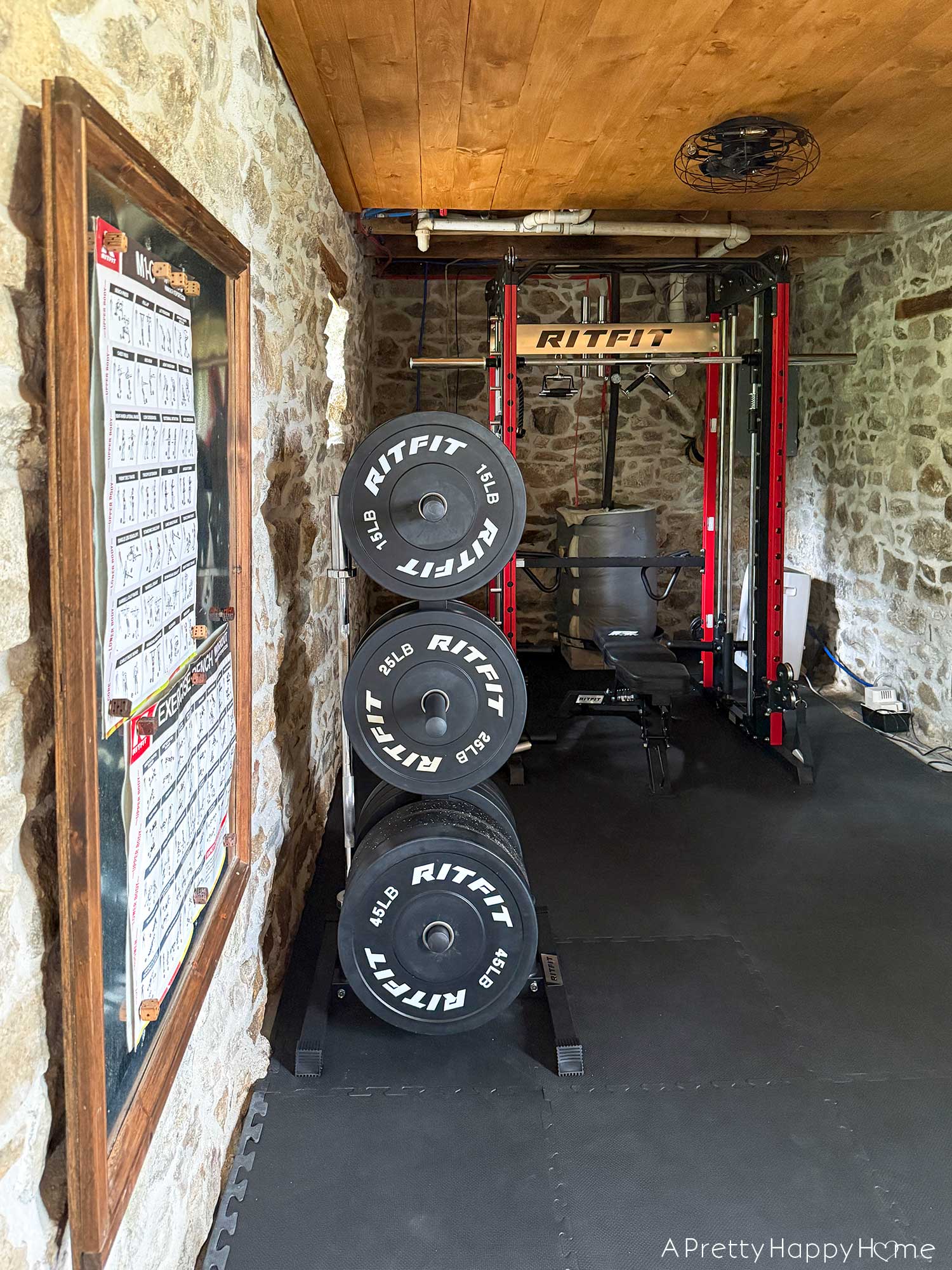 Home gym with fieldstone walls, wood ceiling, and black cushioned floor. There is a red and silver RitFit branded Smith machine in the background with a weight rack with free weights in the foreground. Hanging on the wall is a bulletin board with posters showing weightlifting exercises.