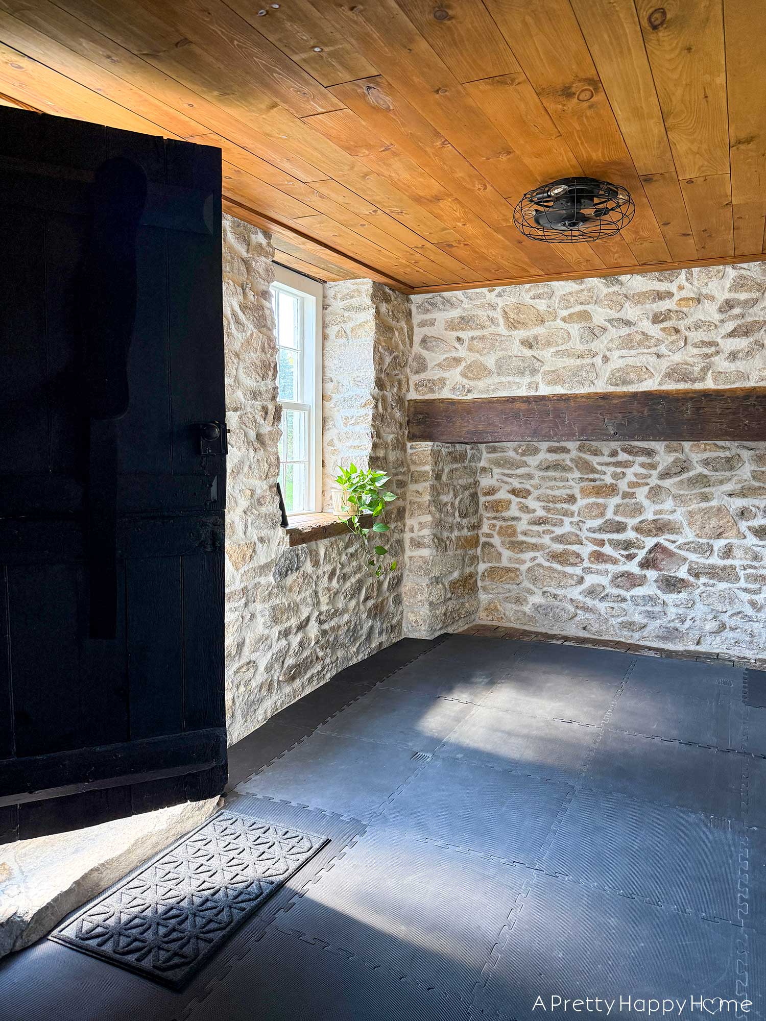 Home gym with fieldstone walls, wood ceiling, and black cushioned floor. Photo doesn't show workout equipment, but shows a half opened black door, a window with a rustic wood windowsill. A pothos plant is sitting on the windowsill in a ceramic planter. There's a large fireplace on the back wall with a rustic wood mantel.