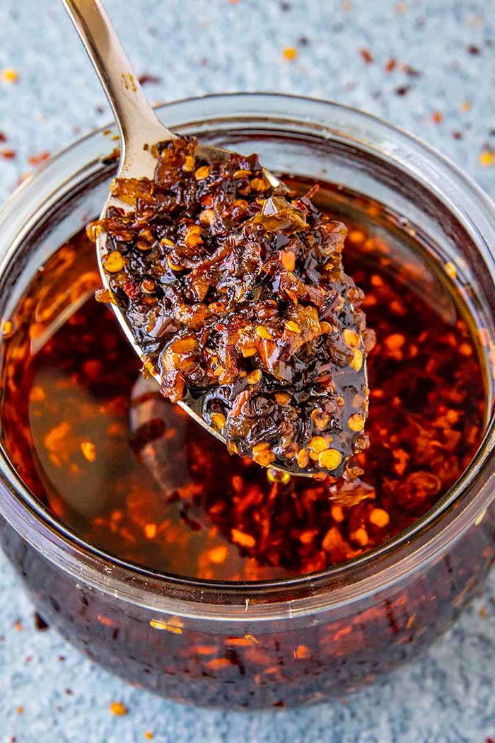 picture of homemade chili crisp with a spoon dipping into a glass jar of chili crisp. the recipe is from chili pepper madness and shown here on the happy list.