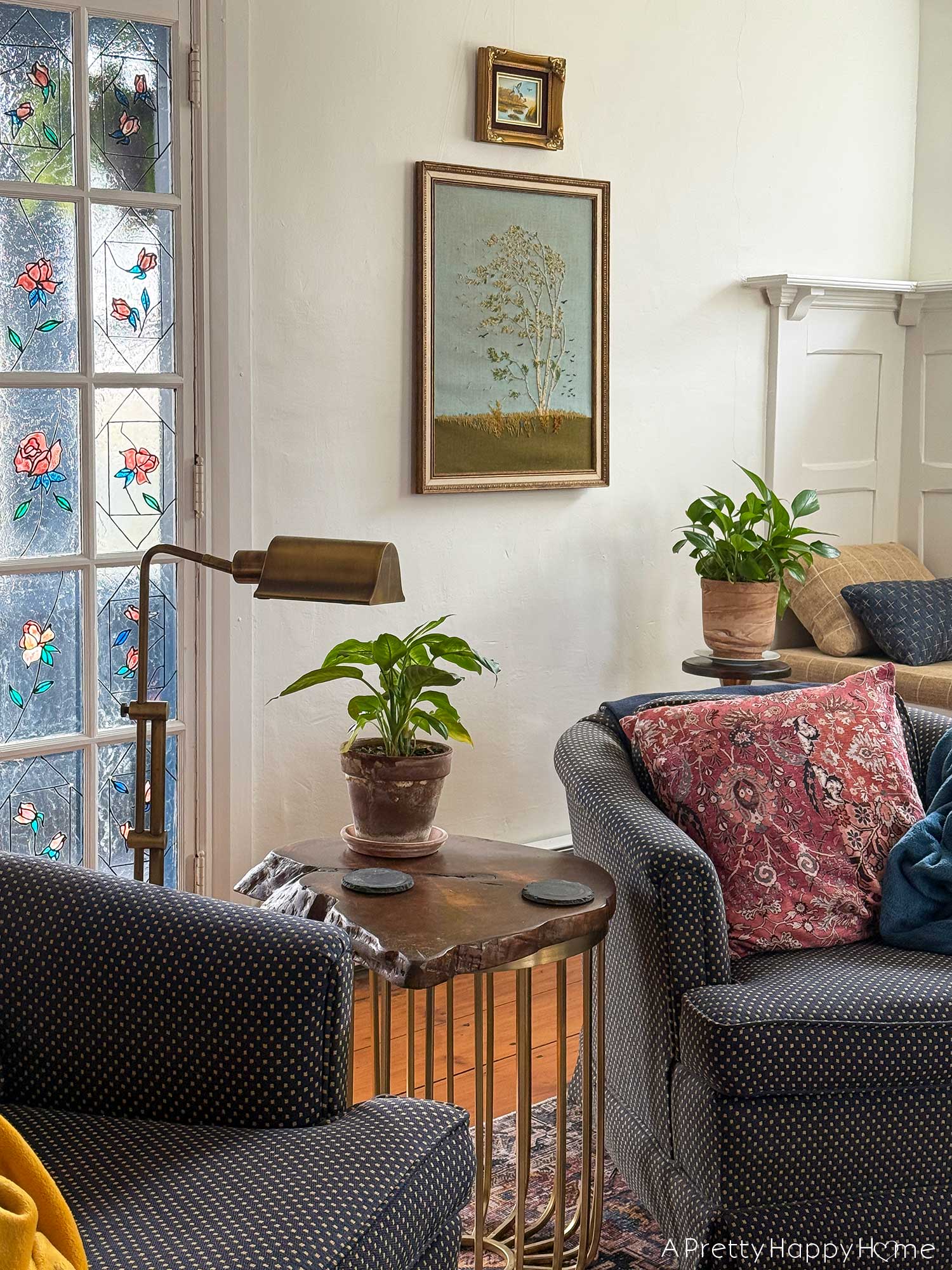 living room vignette focusing on a vintage paragon needlecraft called birch in the wind hanging on the wall. There's a low blue chair sitting in front of the needlepoint. There's also a pothos plant sitting on a wood and brass plant stand in front of the needlepoint, which was thrifted fiber art.