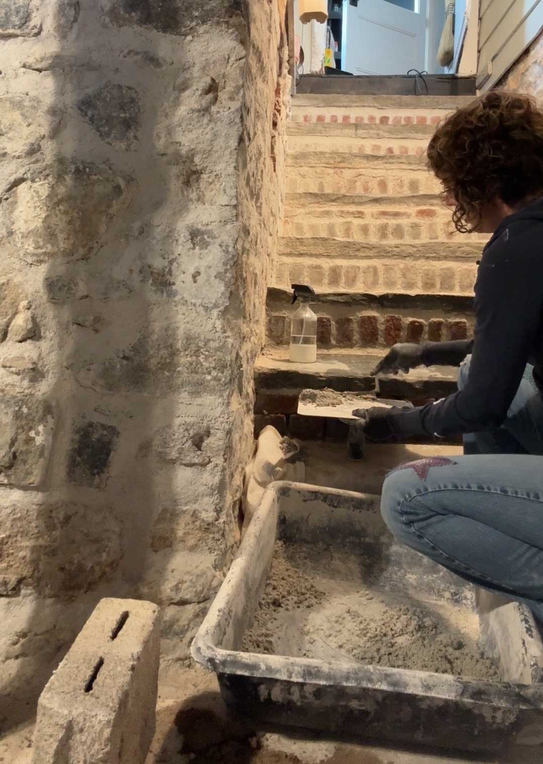 flagstone stairs to a basement with brick risers. the sides of the stairwell are part fieldstone, part brick. A woman with short, curly dark hair is repointing the bricks with lime mortar. She's hiding a trowel and a tray of mortar while a black bin of mortar sits at the bottom of the stairs.