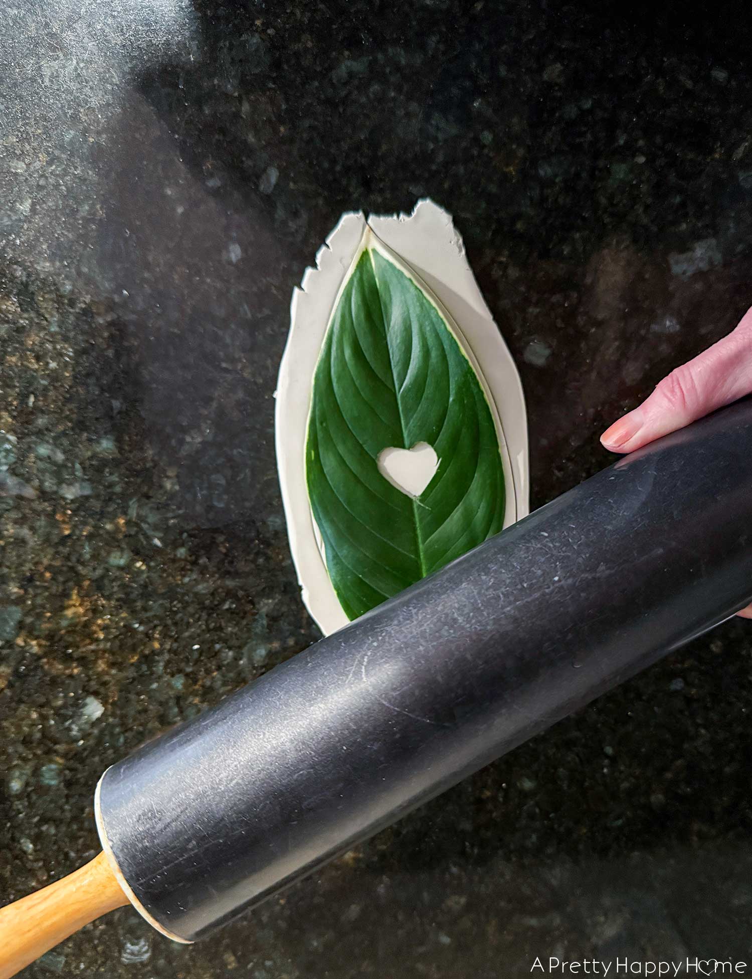 photo a DIY Polymer Clay Leaf Dish with Heart Detail. A leaf with a heart shape cut out of the center is being pressed into fresh oven bake clay with a rolling pin. 