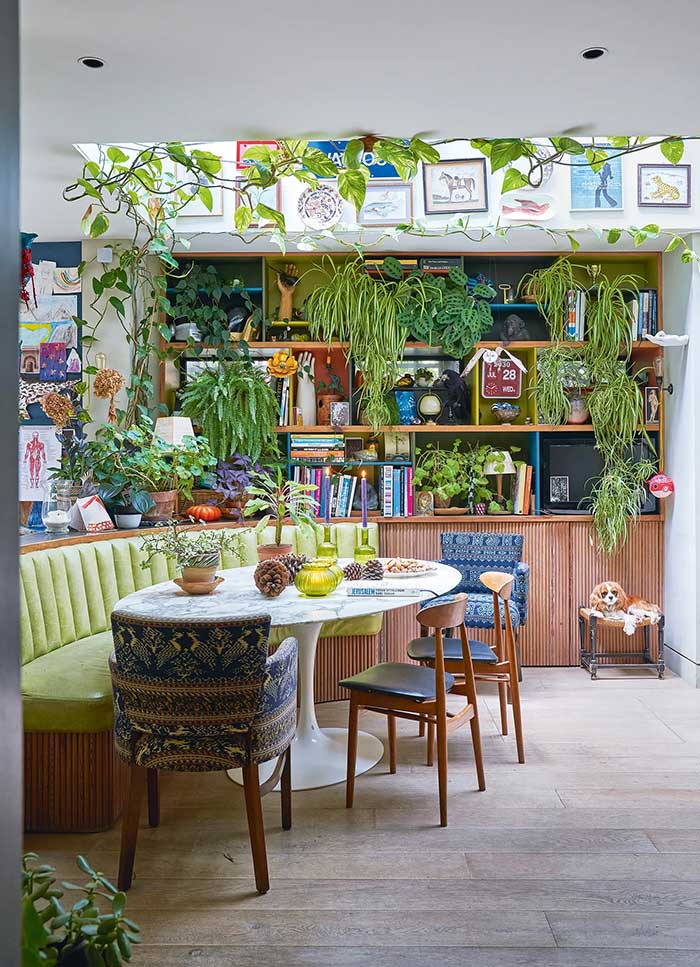 photo credit to Future and James Merrell via homes and gardens. Shows a light wood build in on a wall filled with shelves and cabinets anchoring the bottom. The shelves are filled with plants. the built in connects to a curved banquette with a lime green cushion. There's a table in front of it with more plants. 