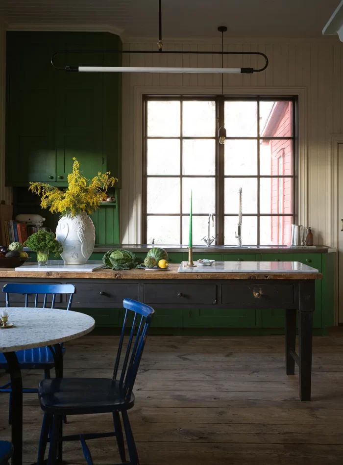 dark cottage core kitchen with large window, green cabinets, and a table as a kitchen island.